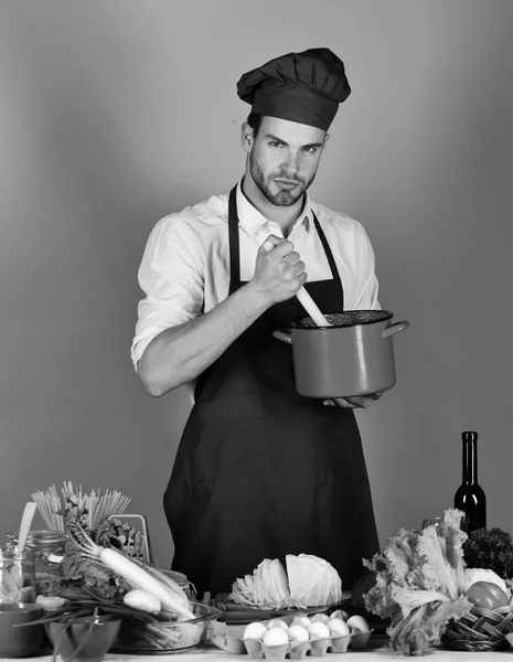 Kock med allvarliga ansikte innehar röd gryta på blå bakgrund. Mannen i kock uniform blandar i kruka med träslev — Stockfoto