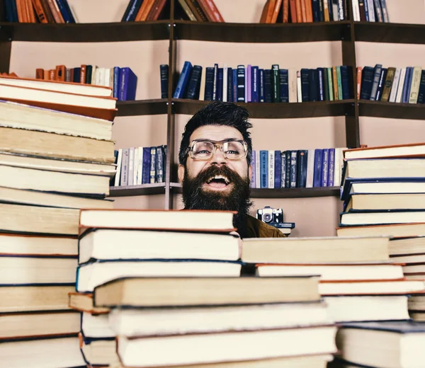 Enseignant ou élève barbu porte des lunettes, s'assoit à table avec des livres, déconcentré. Homme sur le visage excité entre des piles de livres à la bibliothèque, étagères sur fond. Concept de découverte scientifique — Photo