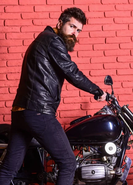 Masculine hobby concept. Man with beard, biker in leather jacket near motor bike in garage, brick wall background. Hipster, brutal biker on serious face in leather jacket sits down on motorcycle