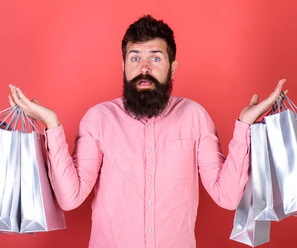 Hombre adicto a las compras con barba larga aislada sobre fondo rojo. Hipster en camisa rosa con barba elegante sosteniendo bolsas de compras de plata. Hombre barbudo con mirada sorprendida gastó todo su dinero a la venta — Foto de Stock