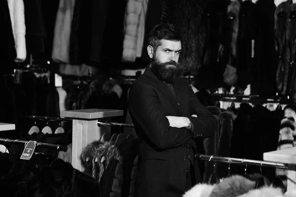Concepto de lujo y ropa elegante. Un tipo con barba cerca de abrigos peludos. El hombre con la cara segura sostiene los brazos cruzados — Foto de Stock