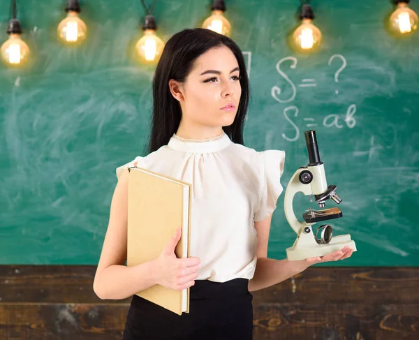 Lady wetenschapper houdt boek en Microscoop, schoolbord op achtergrond, kopie ruimte. Lady in formele slijtage op kalme gezicht in de klas. Biologie concept. Leraar biologie houdt boek en Microscoop — Stockfoto