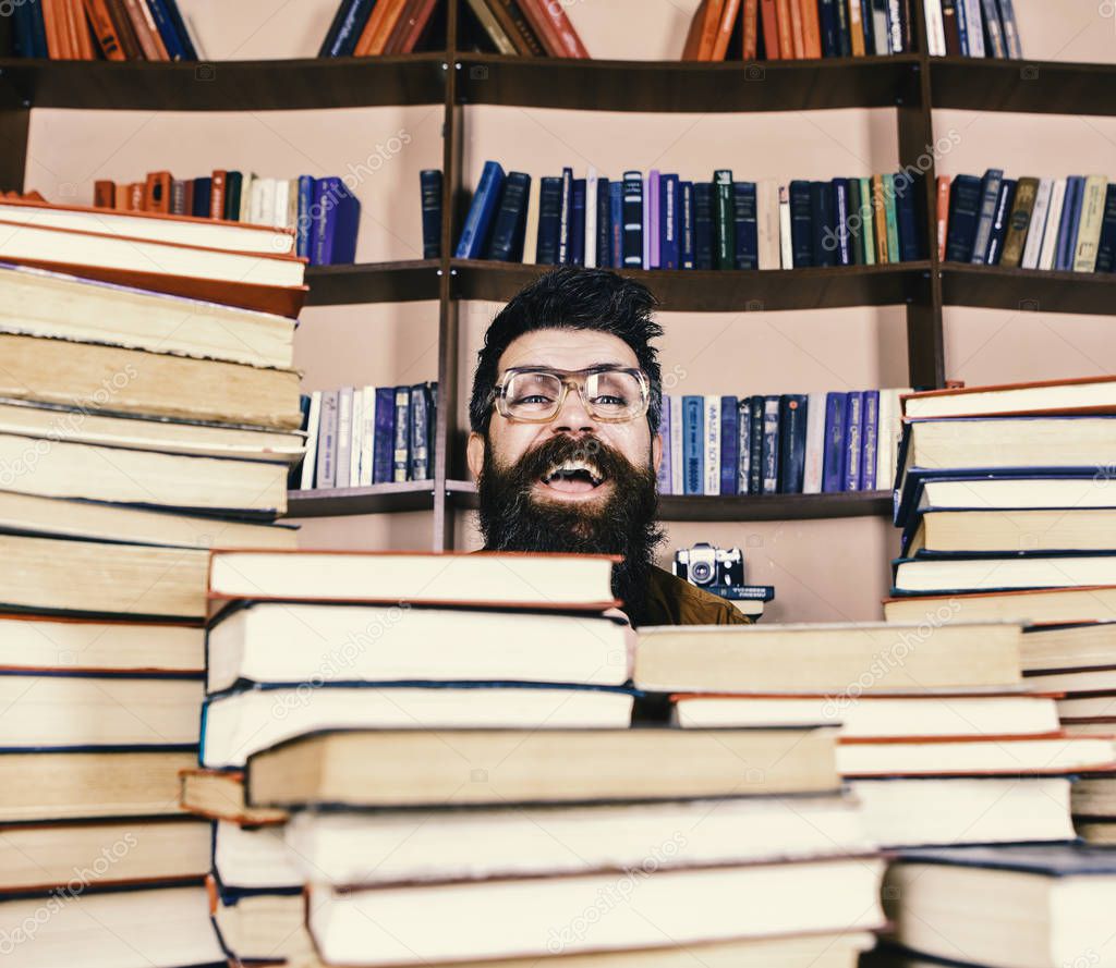 Teacher or student with beard wears eyeglasses, sits at table with books, defocused. Man on excited face between piles of books in library, bookshelves on background. Scientific discovery concept