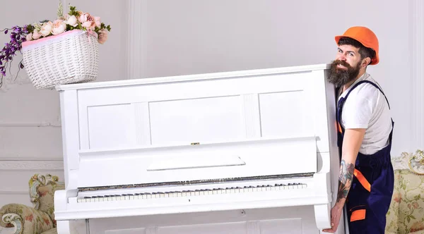 El cargador mueve el instrumento de piano. Servicio de entrega concepto. Hombre con barba, trabajador con mono y casco levanta el piano, fondo blanco. Courier entrega muebles en caso de mudanza, reubicación — Foto de Stock