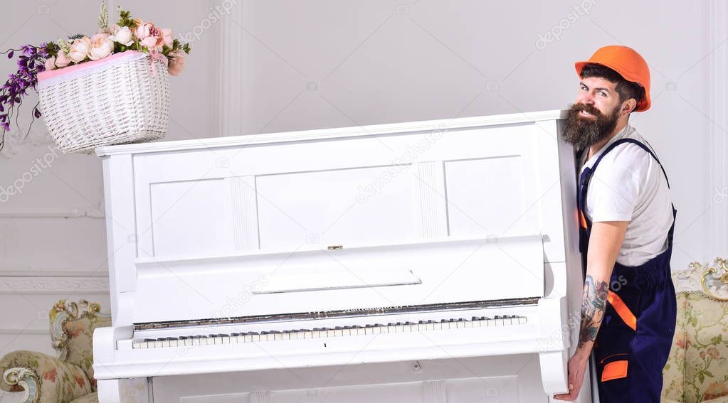 Loader moves piano instrument. Delivery service concept. Man with beard, worker in overalls and helmet lifts up piano, white background. Courier delivers furniture in case of move out, relocation