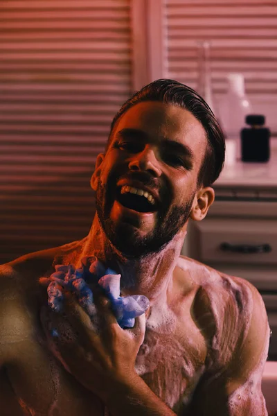 Kaveri pesee kylpyhuoneessa kylpytuotteita taustalla. Mies, jolla on parta ja iloinen naama. Rentoutuminen ja erotiikka käsite: alasti macho pesu kylpyammeessa — kuvapankkivalokuva