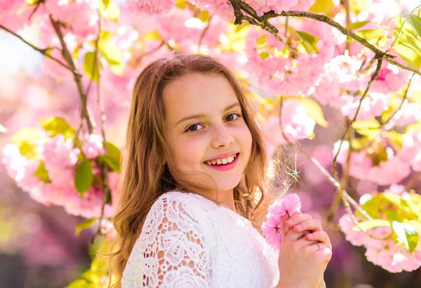 Söta barn njuta av doften av sakura vårdag. Flicka på leende ansikte står nära sakura blommor, oskärpa. Tjej med långt hår utomhus, cherry blossom på bakgrund. Parfym och fragrance-konceptet — Stockfoto