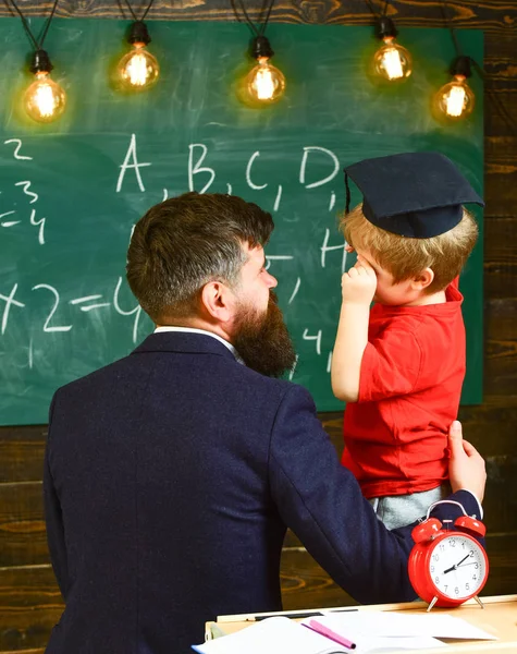 Boy, child in graduate cap discussing scribbles on chalkboard while teacher listening. Teacher with beard, father teaches little son in classroom, chalkboard on background. Prodigy child concept — Stock Photo, Image