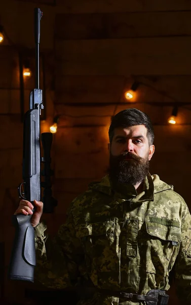 Conceito de guarda-redes. Hunter, um hipster brutal com uma arma na mão pronta para caçar. Macho em rosto estrito na casa dos guarda-redes. Homem com barba usa roupas de camuflagem em fundo interior de madeira — Fotografia de Stock