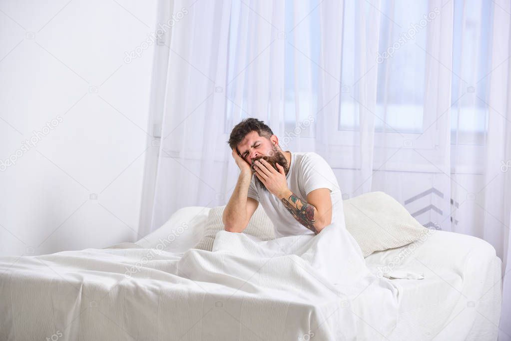 Guy on sleepy tired face yawning. Macho with beard and mustache yawning, relaxing, having nap, rest. Man in shirt sits on bed, white curtains on background. Nap and siesta concept