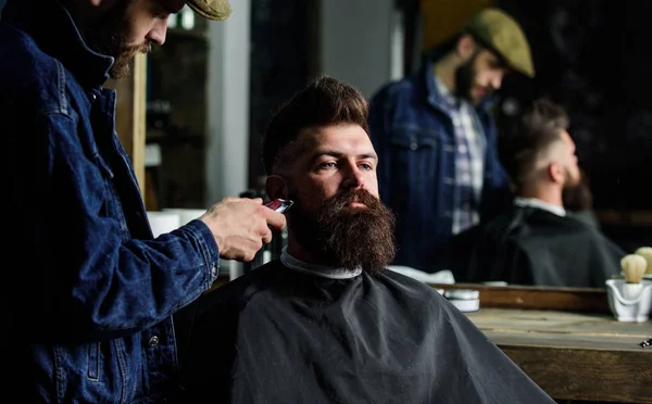 Гіпстер з бородою вкритий обрізанням мису професійним перукарем у стильному перукарні. Концепція вирощування. Перукар зайнятий з доглянутою бородою хіпстерського клієнта, дзеркальна рефлексія на фоні — стокове фото