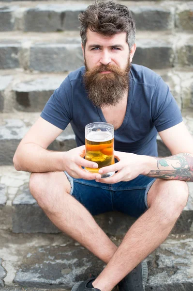 Csípő, hosszú szakállal úgy néz ki, nyugodt. Szakállas csípő tart korsó sör, italok sör szabadtéri. Férfi szakáll és bajusz, arcán nyugodt, köves háttér, defocused. Kézműves sör fogalma — Stock Fotó