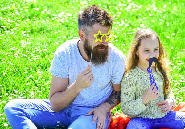 Концепция лучших друзей. Папа и дочь сидят на траве на лужайке, зеленом фоне. Ребенок и отец позируют с очками, усами и атрибутами фотобудки для галстуков. Семья проводит досуг на открытом воздухе — стоковое фото