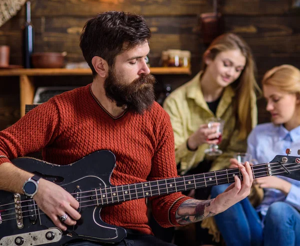 Hombre con barba hipster tocando la guitarra en la fiesta universitaria. Músico ensayando nueva canción. Hombre barbudo entreteniendo a dos chicas rubias. Guitarrista afinando su instrumento. Hombre con cara tranquila componiendo melodía — Foto de Stock