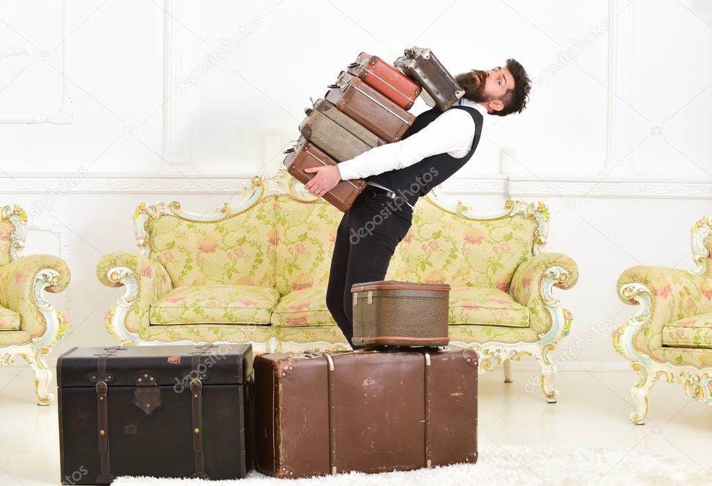 Man with beard and mustache wearing classic suit delivers luggage, luxury white interior background. Macho, elegant porter carries heavy pile of vintage suitcases. Butler and service concept