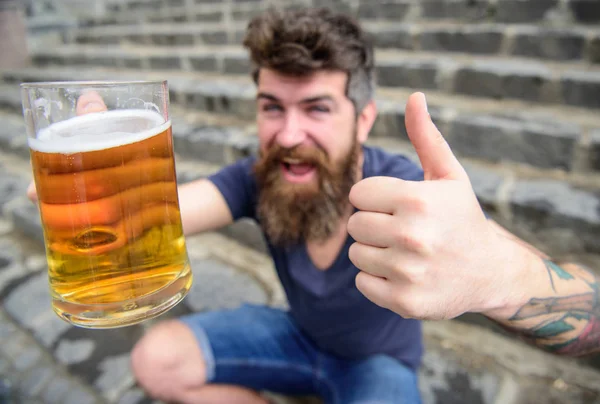 Hipster en la cara alegre bebe cerveza. Concepto de cerveza de barril. Un tipo descansando con cerveza, enfoque selectivo. Hombre con barba y bigote sostiene el vaso con cerveza y muestra el pulgar hacia arriba, escaleras de piedra de fondo — Foto de Stock