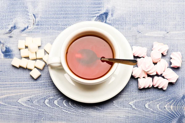 Mugg med svart te och godis. Te paus koncept. Dryck och sötsaker förberett som efterrätt. Mugg fylld med svart bryggt te, sked och högen av marshmallow och raffinerat socker på blå trä bakgrund — Stockfoto