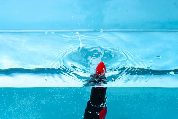 Resistencia al agua concepto cosmético. Lápiz labial bajo el agua con burbujas transparentes y salpicaduras de líquido. Lápiz labial rojo resistente al agua caer en pur, agua transparente, fondo azul —  Fotos de Stock