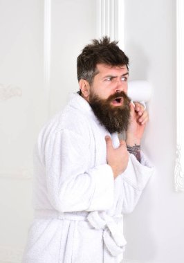Man with beard and mustache eavesdrops using mug near wall. Hipster in bathrobe on concentrated face secretly listen conversation. Secret and spy concept. Man in white interior spying, eavesdropping clipart