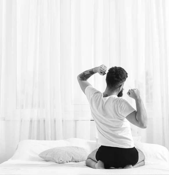 El hombre de camisa se sienta en la cama, cortinas blancas en el fondo. Tipo estirando los brazos, lleno de energía en la mañana, vista trasera. Concepto matutino perfecto. Macho en ropa interior estirándose, relajándose después de la siesta, descansar —  Fotos de Stock