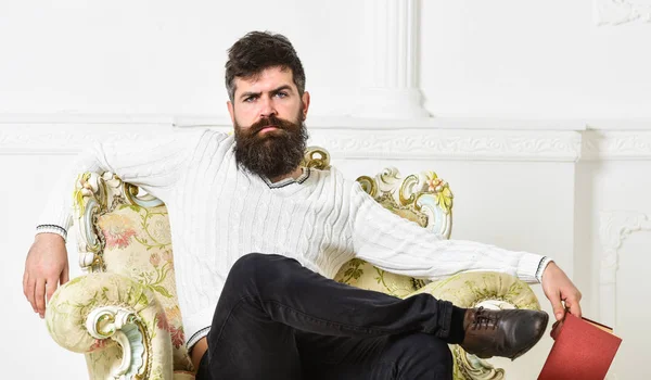 Hombre con barba y bigote se sienta en el sillón, sostiene libro, fondo blanco de la pared. Conocedor de cara reflexiva terminado libro de lectura. Conocedor del concepto de literatura. Chico pensando en literatura —  Fotos de Stock