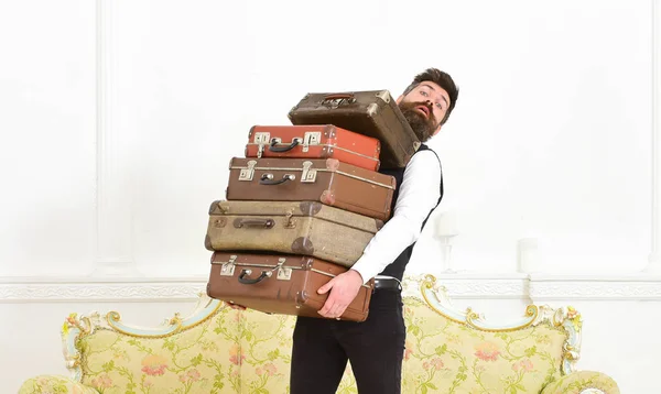 Man with beard and mustache wearing classic suit delivers luggage, luxury white interior background. Butler and service concept. Macho elegant on surprised face carries pile of vintage suitcases