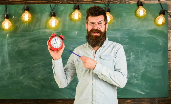 Skäggiga hipster innehar klocka, svarta tavlan på bakgrund. . Lärare i glasögon håller väckarklocka och penna. Disciplin-konceptet. — Stockfoto