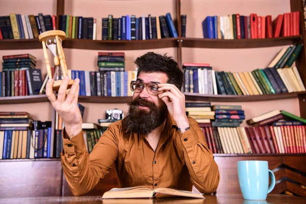 Professor ou estudante com barba estudando na biblioteca. O cientista com óculos senta-se à mesa e olha para a ampulheta. Homem na cara ocupada lendo livro, estantes de livros no fundo. Lição e conceito de tempo — Fotografia de Stock