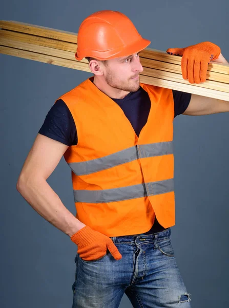 Concepto de trabajador resistente. Carpintero, carpintero, obrero, constructor en la cara ocupada lleva vigas de madera en el hombro. Hombre en casco, sombrero duro y guantes de protección sostiene la viga de madera, fondo gris —  Fotos de Stock