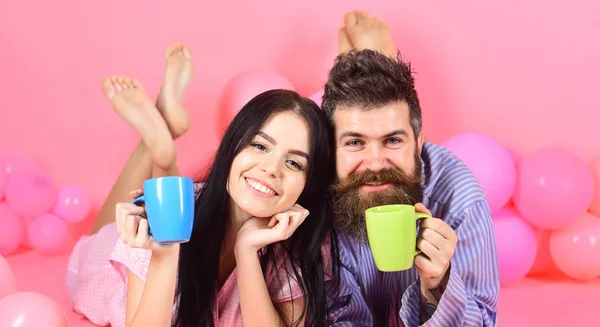 Hombre y mujer en rostros sonrientes yacían, fondo rosa. Pareja enamorada bebe café en la cama. Pareja relajarse en la mañana con café. Concepto de tradición familiar. Hombre y mujer en ropas domésticas, pijamas — Foto de Stock