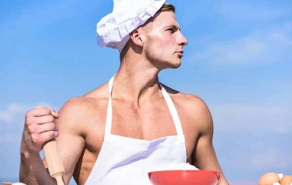 L'uomo sul viso sicuro indossa cappello da cucina e grembiule, cielo sullo sfondo. Concetto di cucina. Cuoco cuoco preparare la pasta per la cottura. Cuoco o chef con sexy spalle muscolose e petto tiene mattarello — Foto Stock