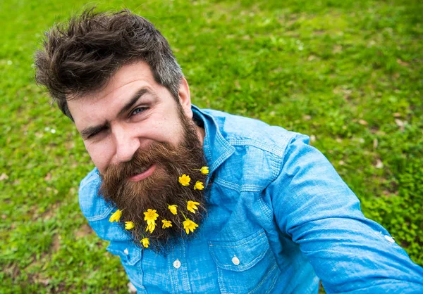 Yüz göz kırpıyor üzerinde hipster defocused çimenlerin üzerinde oturur. Sakallı bahar, yeşil çayır arka plan sahiptir. Küçük kırlangıçotu çiçeklerle selfie fotoğrafta sakallı adam. Bahar sezonu kavramı
