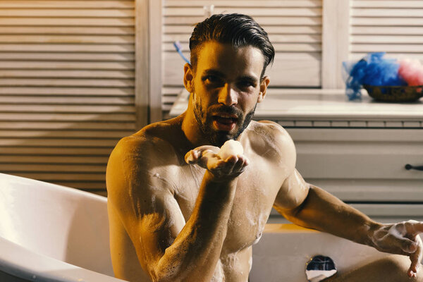 Человек с бородой и очаровательным лицом, выдувающим пену из ладони. Парень в ванной, покрытый пеной
