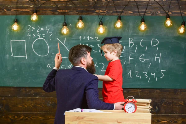有益的谈话概念。老师带着胡子, 父亲在教室里教小儿子, 黑板上的背景。孩子在研究生盖帽听力老师, 黑板在背景, 后方看法 — 图库照片