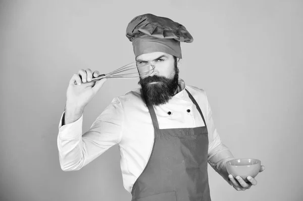 Chef-kok met rode plaat en zweepslagen gebruiksvoorwerp. Man of hipster met baard houdt keukengerei op rode achtergrond. Keuken hulpmiddelen concept. Koken met ernstige gezicht in Bourgondië hoed — Stockfoto