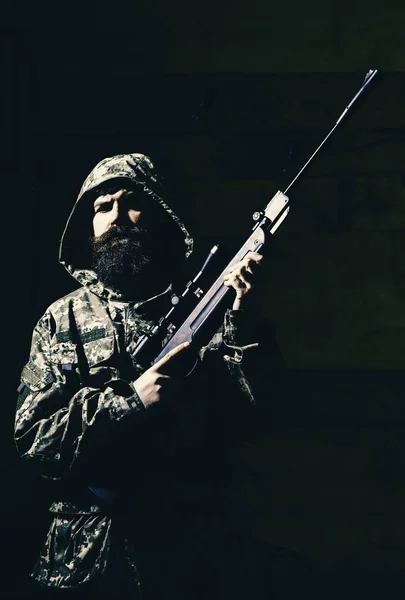 Hunter, brutal con arma prepara rifle para cazar. Concepto de cazador. Macho en cara estricta en la casa de los guardabosques. Hombre con barba lleva ropa con capucha camuflaje, fondo interior de madera . —  Fotos de Stock