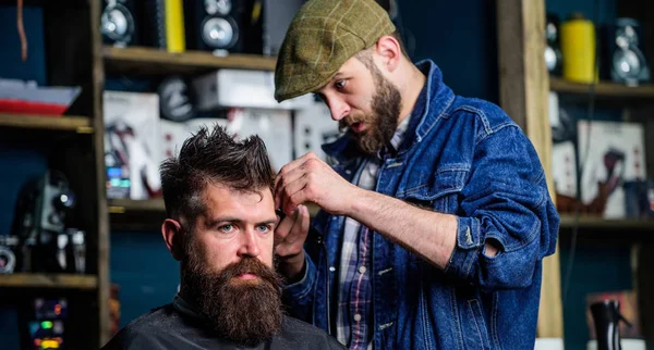 Barber in denim jacket busy with trimming hipster, barbershop background. Client with beard and mustache covered with cape. Image making concept. Barber work on haircut with hair clipper equipment — Stock Photo, Image
