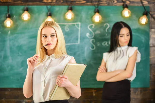 Envy and competition concept. Girl jealous of success of classmate in classroom, chalkboard on background. Woman with book and eyeglasses looks dreamy while student on background looks jealous — Stock Photo, Image