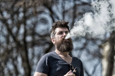 Elektronik Sigara kavramı. Uzun sakallı ve bulutlar duman görünüyor rahat olan adam. Sakal ve bıyık sakin yüzündeki defocused arka plan, dallarda olan adam. Sakallı adam vape güneşli günde içiyor.