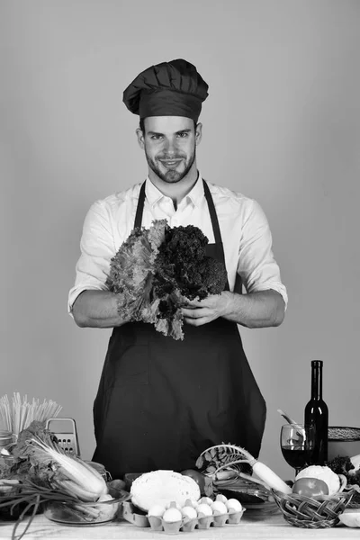 Mannen i kock hatt och förkläde med sallad. Cook fungerar i köket med grönsaker och verktyg. — Stockfoto