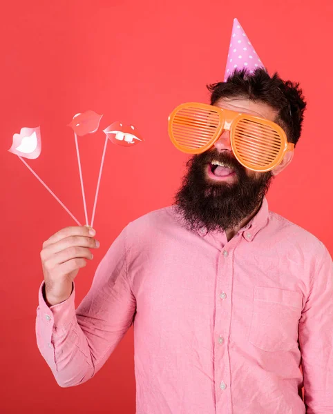 Emotionale Vielfalt. Mann mit Bart auf fröhlichem Gesicht hält lächelnde Lippen auf Stöcken, roter Hintergrund. Mann mit Partyhut feiert, posiert mit Fotorequisiten. Hipster mit Riesensonnenbrille feiern — Stockfoto