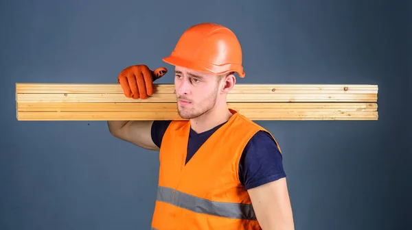 Carpintero, carpintero, fuerte constructor en la cara seria lleva la viga de madera en el hombro. Hombre con casco, sombrero duro y guantes de protección sostiene la viga de madera, fondo gris. Concepto de materiales de madera —  Fotos de Stock