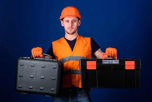 Homem de capacete, chapéu duro segura caixa de ferramentas e mala com ferramentas, fundo azul. Caixa de ferramentas e conceito de equipamento. Trabalhador, reparador, reparador, construtor no equipamento pensativo da escolha da cara para o trabalho — Fotografia de Stock