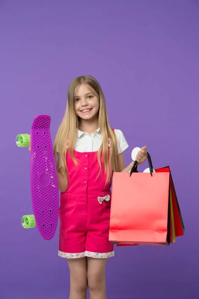 Chica feliz mantenga penny board y bolsas de compras sobre fondo violeta. Sonrisa infantil con paquetes y monopatín. Patinadora sonriendo con bolsas de papel. Compras y venta. Deporte y hobby activo —  Fotos de Stock