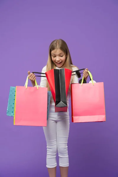 Kind kijk in boodschappentassen op violette achtergrond. Weinig shopaholic glimlach verrast met papieren zakken. Jeugd. Winkelen en verkoop. Shopper meisje met paperbags. Wat een verrassing. Grote verkoop en winkelen — Stockfoto