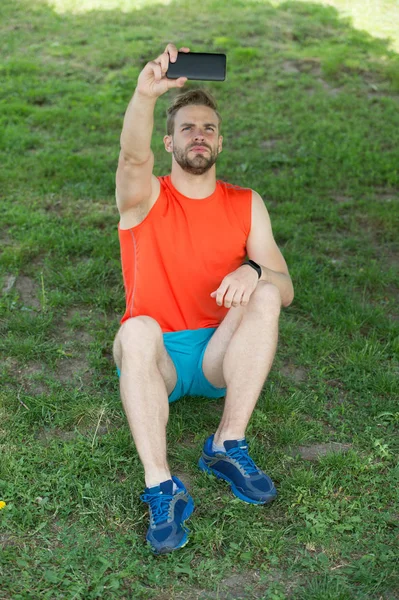 Yeni teknoloji ve modern yaşam. Maço sit smartphone ile yeşil çimenlerin üzerinde. Yaz etkinliği ve spor. Adam olun selfie cep telefonu ile temiz hava. Sporcu eğitim sonra sakin ol. Spor ve fitness