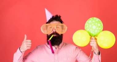 Başparmak gösterilmesini sakallı adam. Parti düdük üfleme trendy sakallı adam. Kırmızı bir arka plan üzerinde renkli balonlarla hippi. Sakallı adam partide kutlama kavramı eğleniyor Doğum günü şapkalı