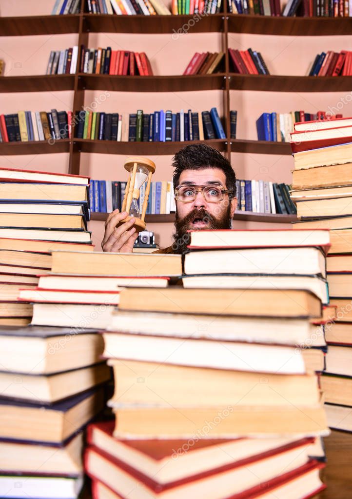 Time flow concept. Man on surprised face holds hourglass while studying, bookshelves on background. Teacher or student with beard studying in library. Man, scientist in glasses looks at hourglass