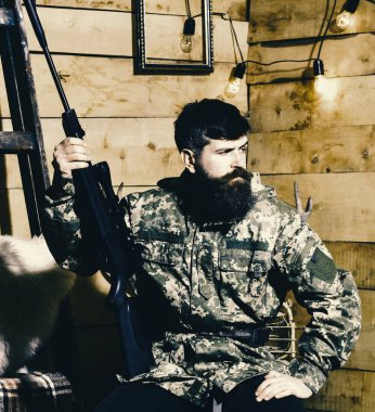 Sakallı adam ahşap iç arka planda kamuflaj elbiseler giyer. Avcı, av için hazır elinde tabanca ile acımasız hipster. Hakem kavramı. Maço gamekeepers evinde sıkı yüzünde.
