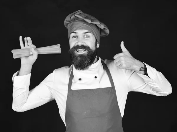 Homme ou hipster à la barbe tient des macaronis sur fond noir. Chef avec spaghettis montre pouces vers le haut. Cuisiner la nourriture italienne — Photo